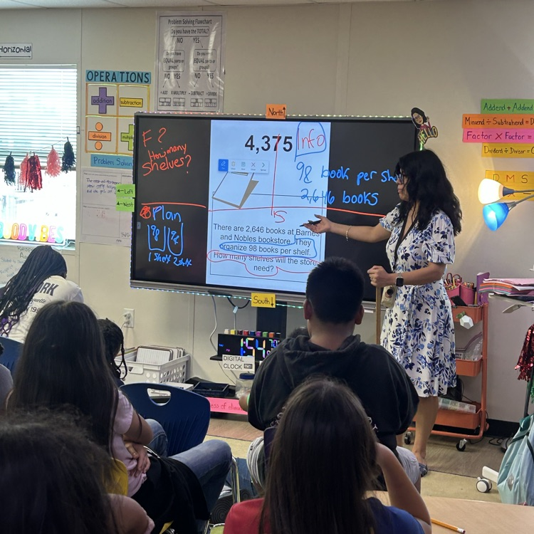 teacher in white dress completing problem-solving board with students