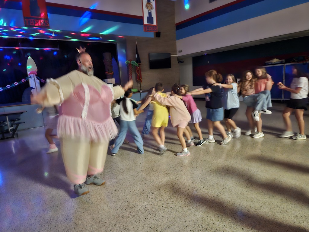 students dancing in a conga line with an adult dancing in an inflatable ballerina costume in the cafeteria with a disco light 