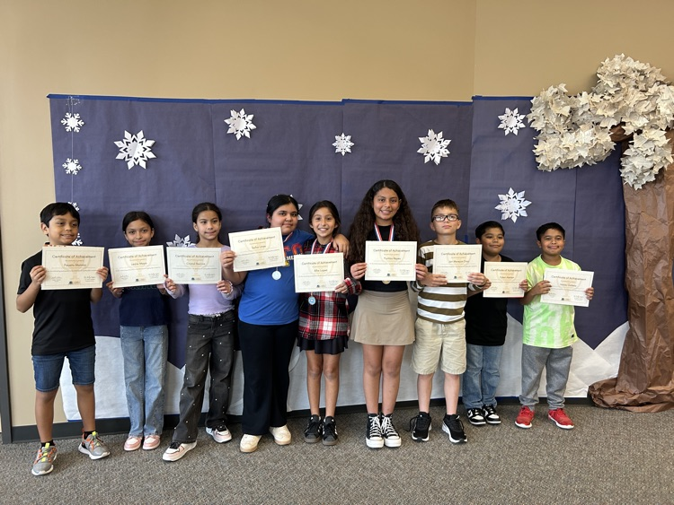 Nine students stand in front of a winter background holding their spelling bee certificates