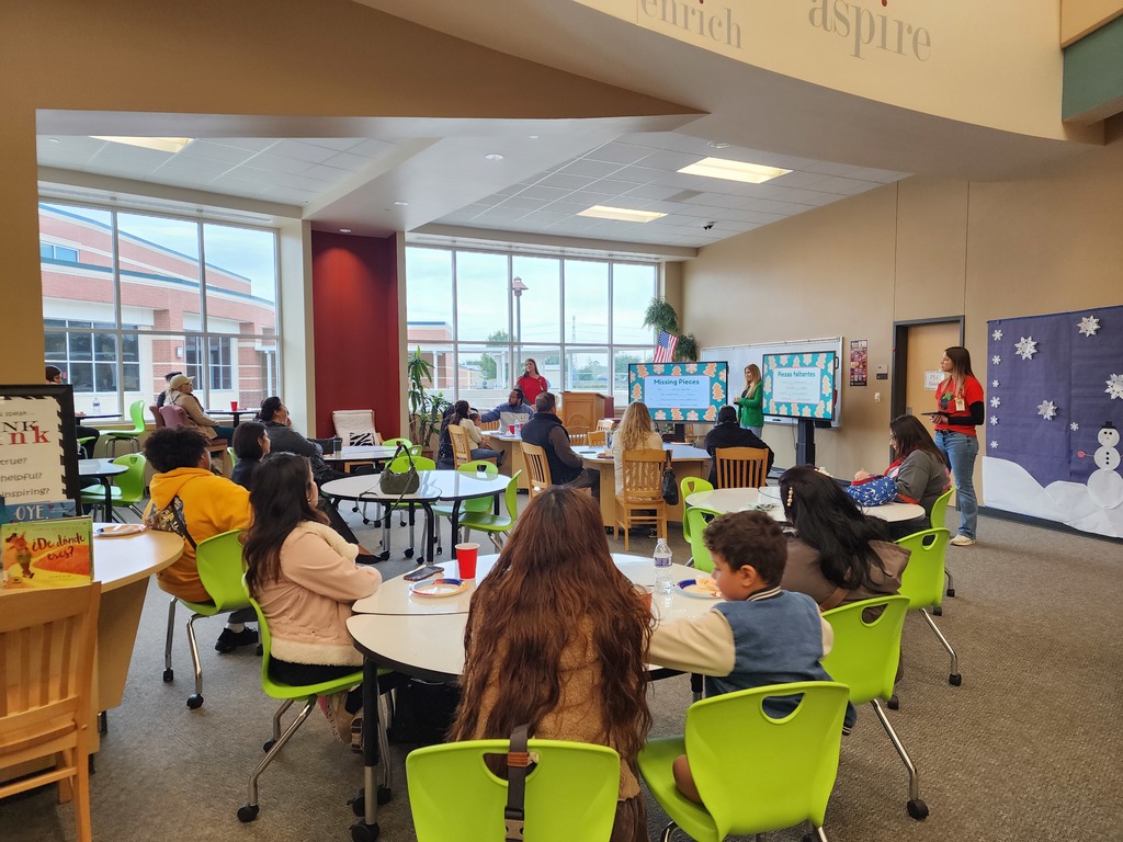 Families seated at tables listening to three presenters standing in the front of the library.