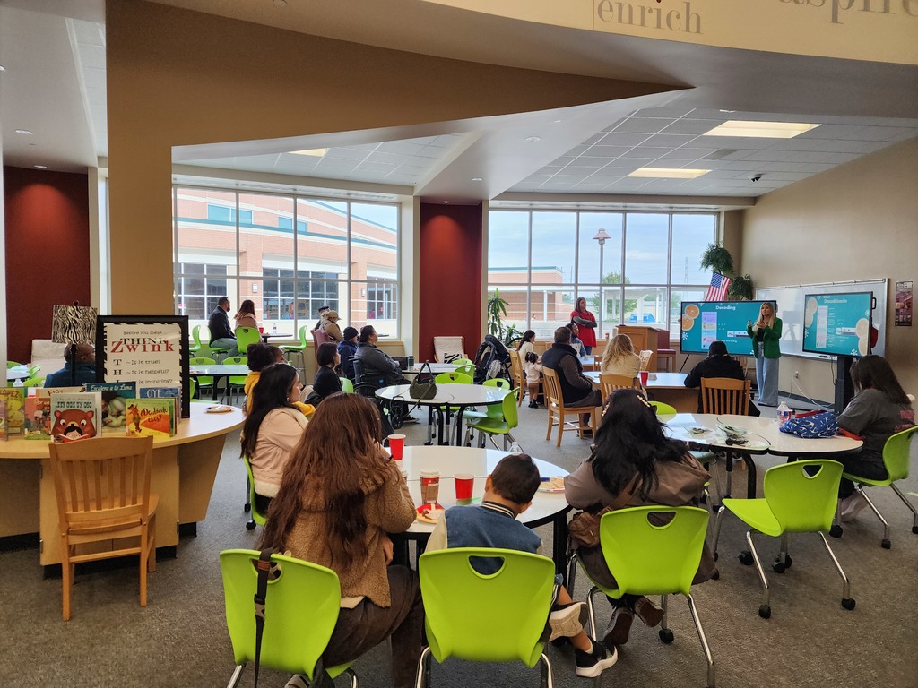 Families seated at tables listening to two presenters standing in the front of the library.