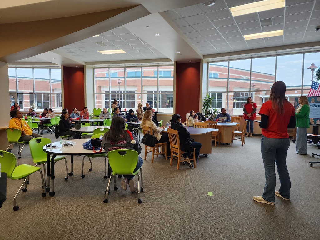 Families seated at tables listening to three presenters standing in the front of the library.