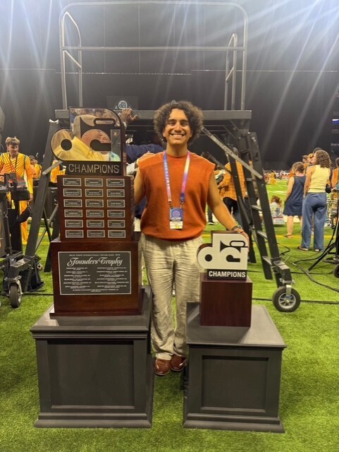 Person in orange shirt stands with two trophies on a field. Background includes people in yellow uniforms and a stage.