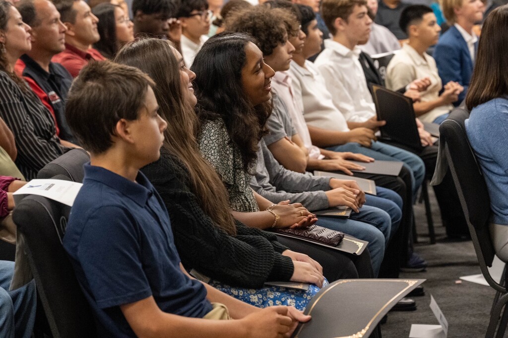 A group of students are seated in a lecture hall, some using laptops. They are focused on the front, where a presentation is likely taking place.