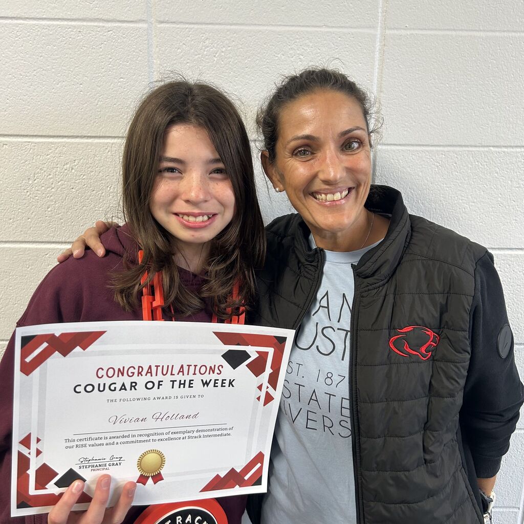 Two people smile in front of a white wall, one holding a certificate for "Cougar of the Week."