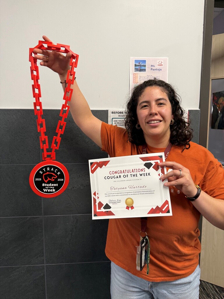 Person in orange shirt and jeans holding a red necklace and certificate with "Cougar of the Week" title.