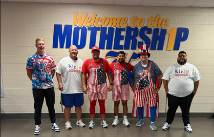 Five men in red, white, and blue outfits pose in front of a wall with "Welcome to the MOTHERSHIP" banner.