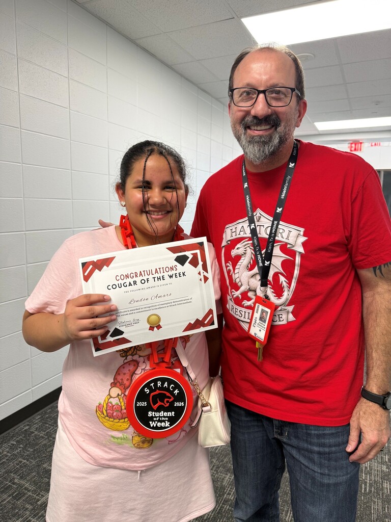A smiling woman holding a certificate stands next to a man with a lanyard, both in a hallway.