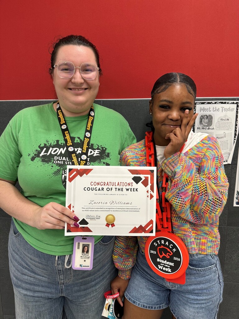 Two women in a school corridor hold a certificate, one wearing green and the other in a multicolored sweater.