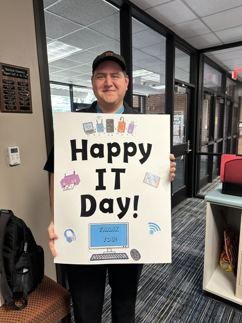 A person stands in a hallway holding a sign that reads "Happy IT Day!" with a smiling face.