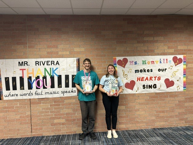 Two people stand before a brick wall with posters. One reads "Mr. Rivera Thank You," and the other says "Ms. Konville makes our hearts sing."
