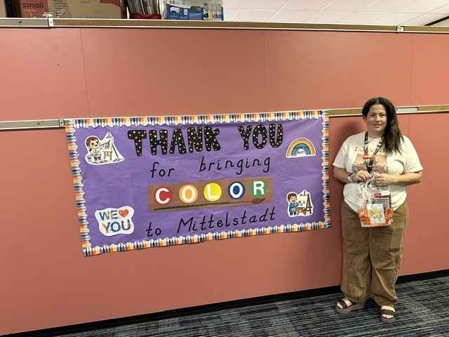 Person stands by a sign with "Thank You for bringing color to Mittelstadt". The sign has a rainbow and colorful letters.