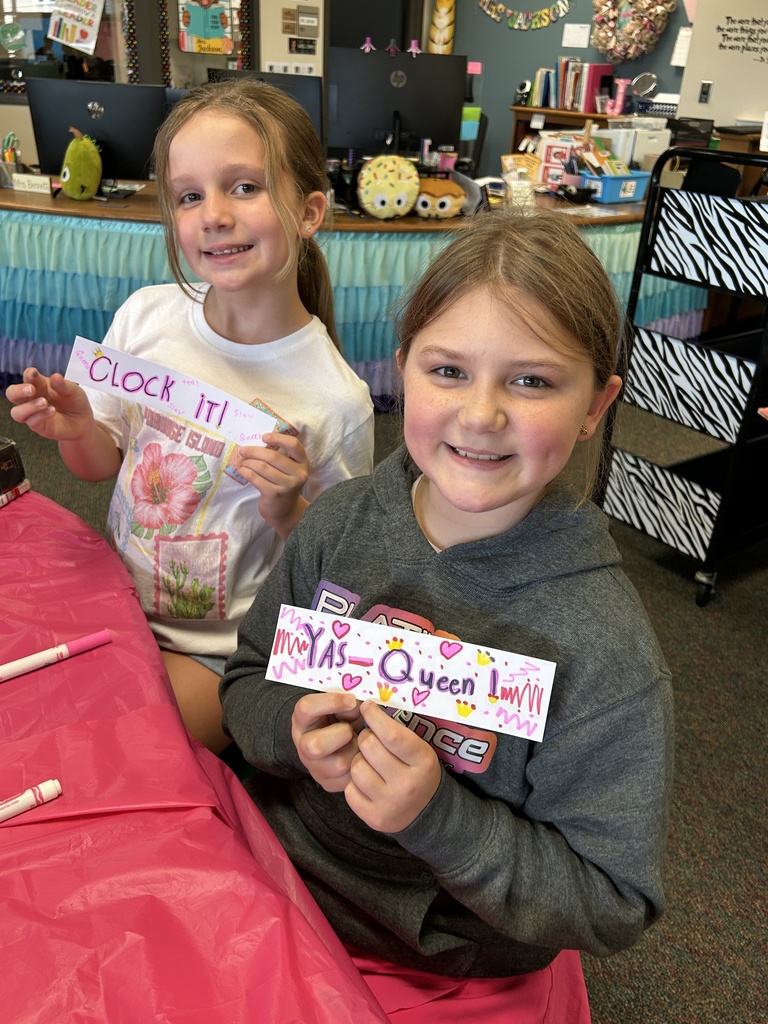 Two girls seated at a table, one with a sign that says "Clock it!" and the other with a sign "XS-Queen Love."