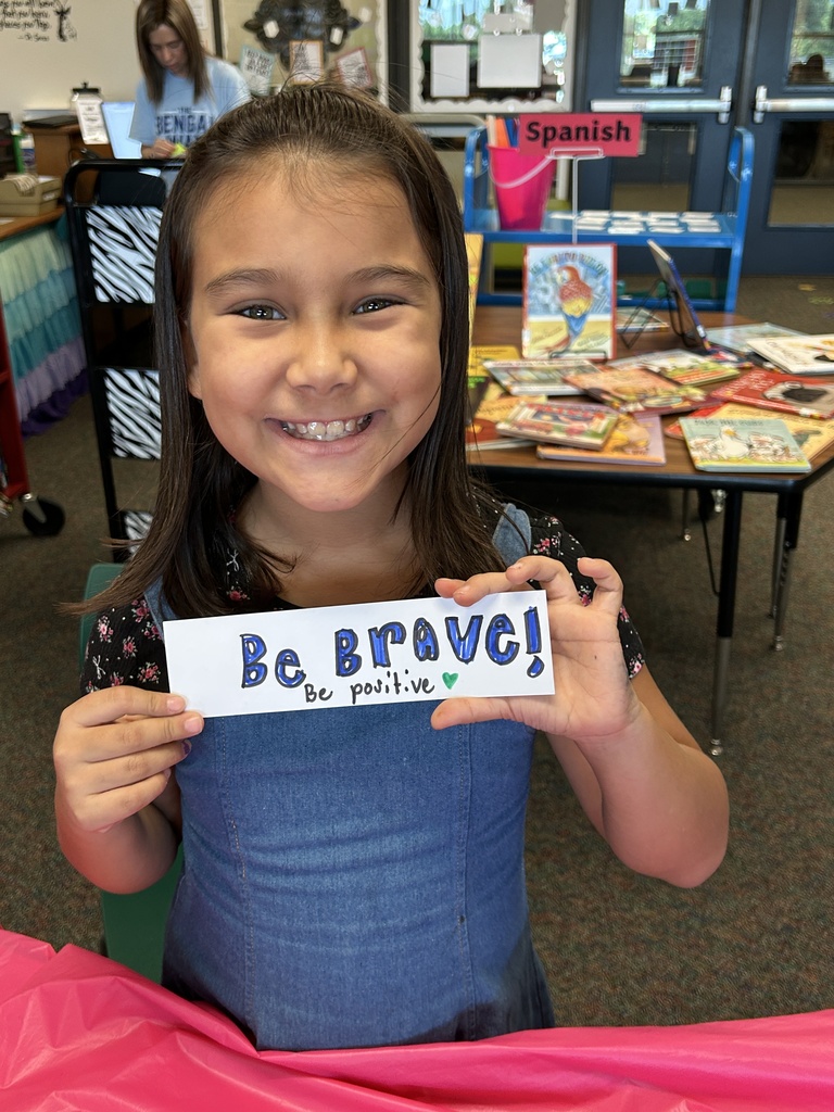 A girl in a blue dress holds a sign that reads "Be Brave!" She is smiling in a classroom.