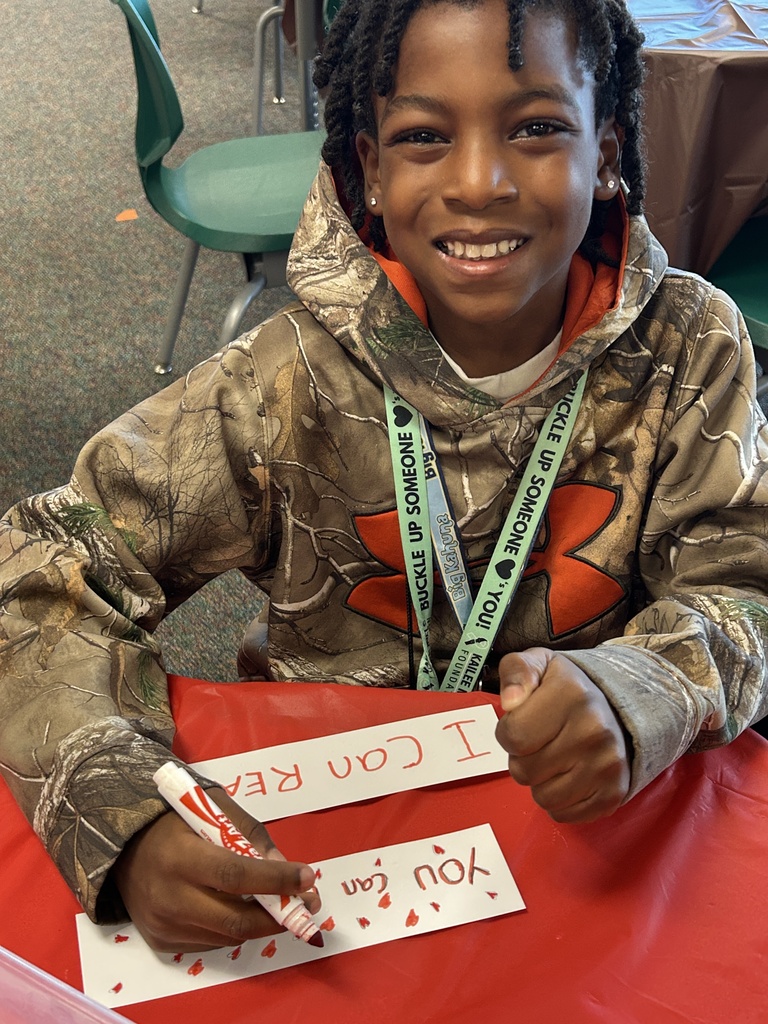 A child sitting at a table holding a marker and writing on paper with red letters.