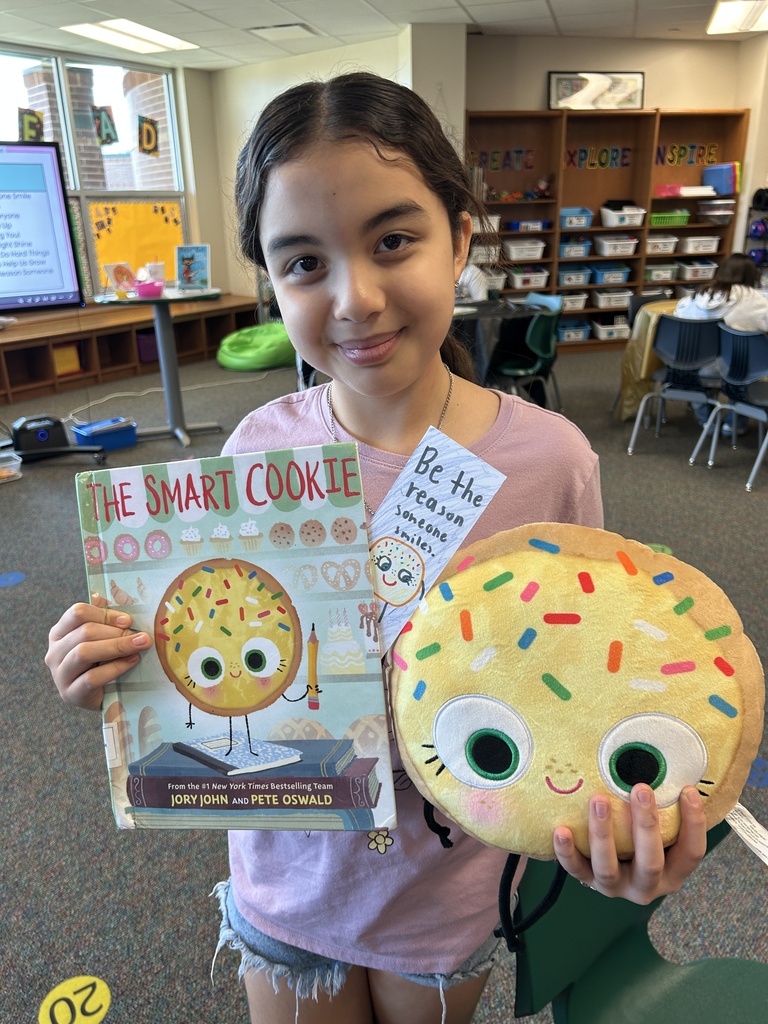 A young girl holding a book titled "The Smart Cookie" and a cookie plush toy. Behind her, a classroom with shelves, a table, and chairs.