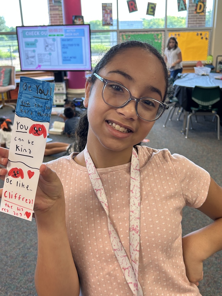 A girl with glasses smiles while holding a paper with "You can be kind" in a classroom setting.