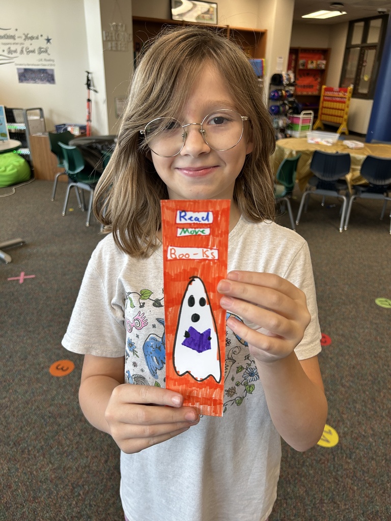 A young girl in a classroom holds a candy wrapper featuring a ghost. She wears glasses and a white t-shirt.