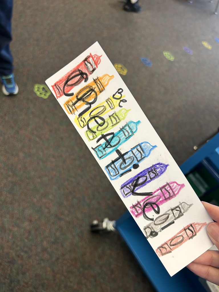 A hand holds a bookmark with colorful markers and text. Behind the person, a floor with multicolored shapes and a blue storage box.