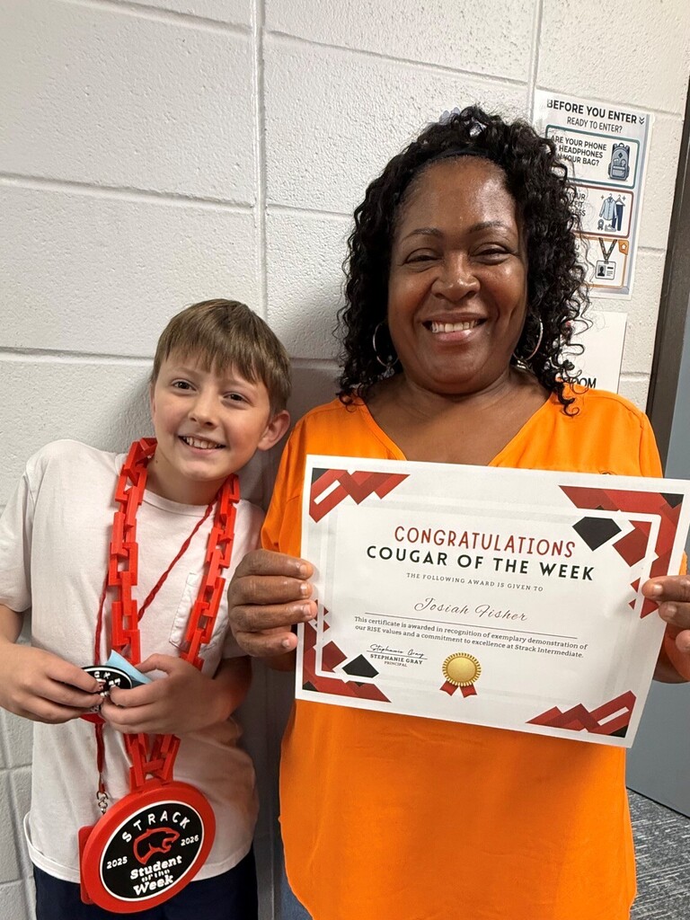 A woman and a boy stand together, both smiling. The woman holds a certificate and a medal, the boy wears a medal. Behind them is a white wall with posters.