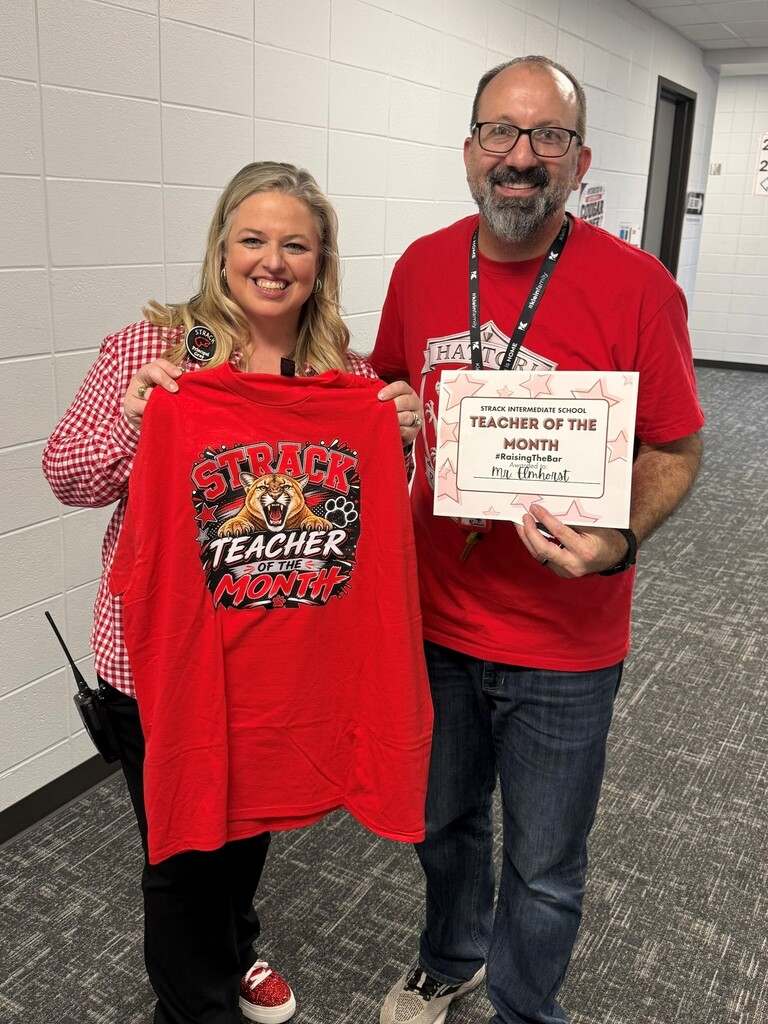 Two people in red shirts stand in a hallway; one holds a red shirt and a certificate, while the other holds a certificate.