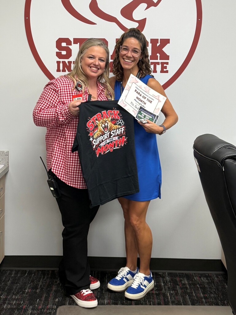 Two women stand in front of a wall with a logo, one holding a certificate and a t-shirt.