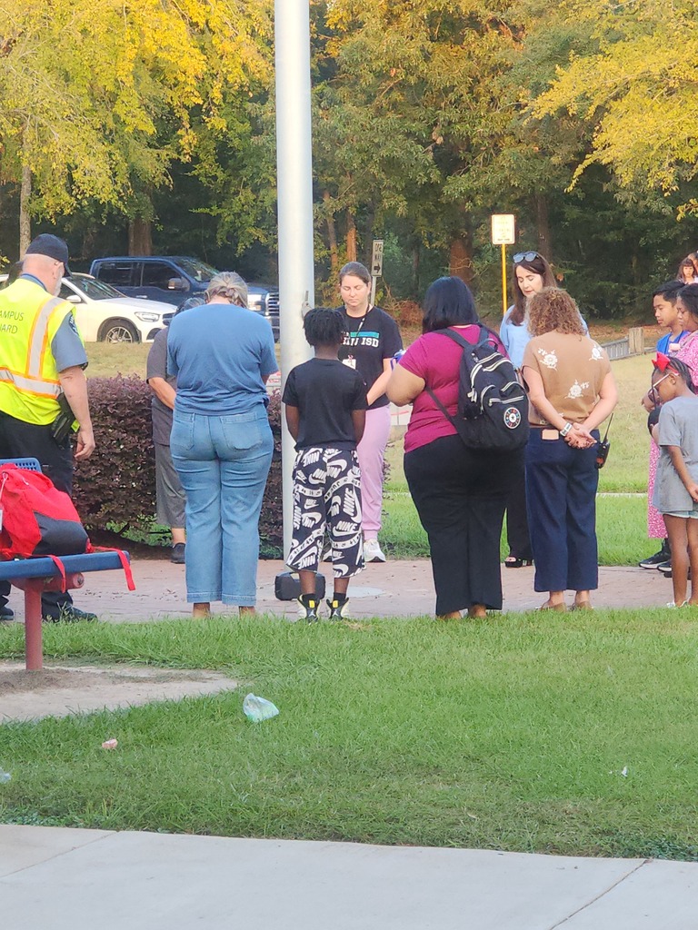 Group of people gathered on a grassy park area. A man with a yellow vest stands nearby. Trees and bushes provide a natural background.