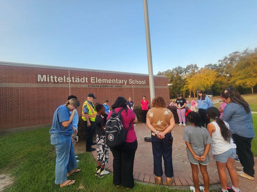 People stand in a circle around a flagpole outside a school. Some wear backpacks, and others hold hands.