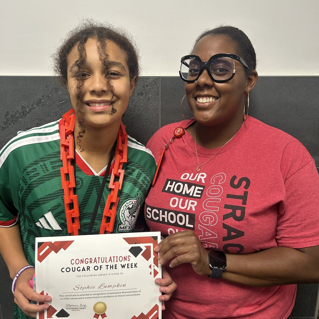 Two women smiling. One holds a certificate. Behind them, a black wall with "OUR HOME OUR SCHOOL STRA" written on it.
