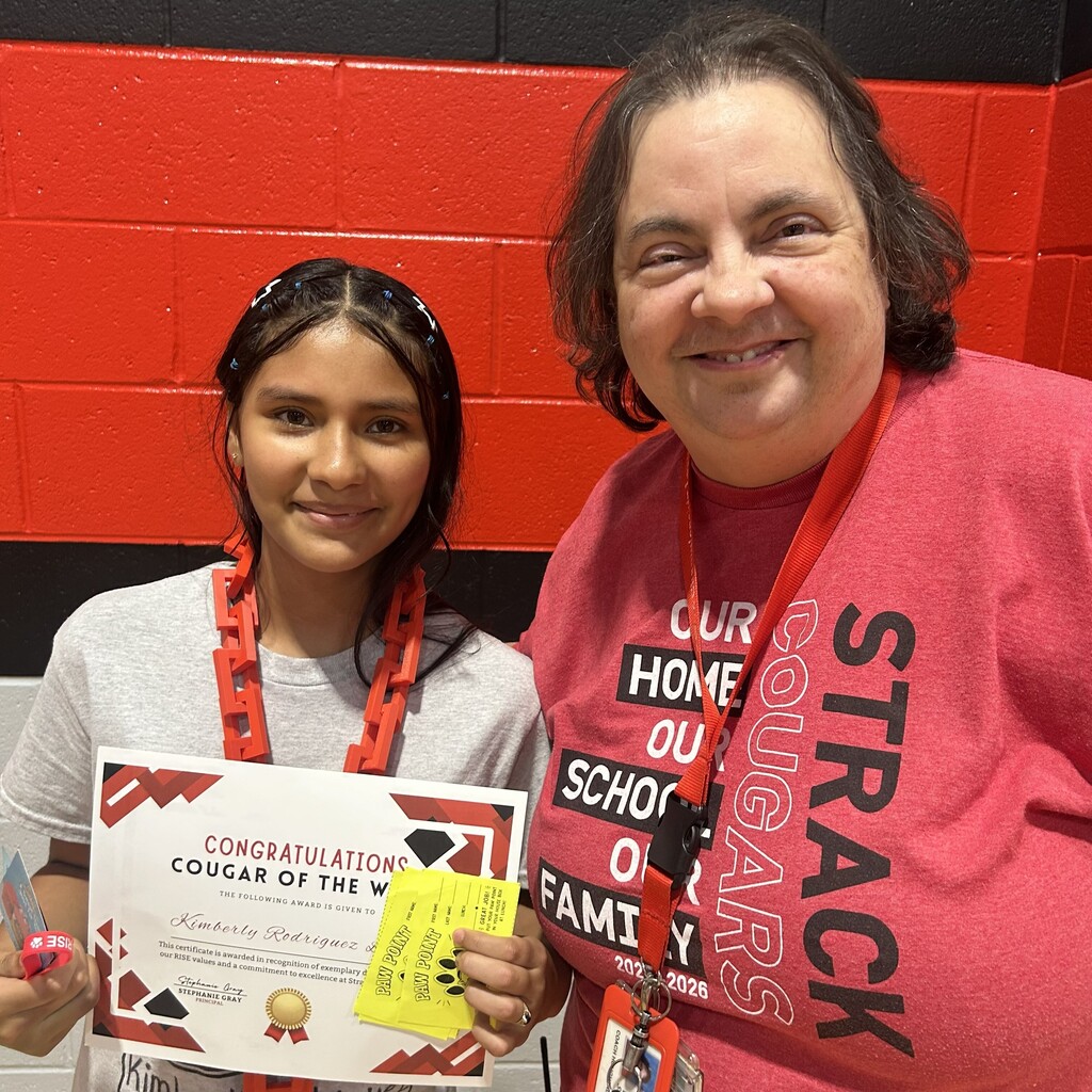 Two people smile, one holding a certificate, standing in front of a red brick wall.