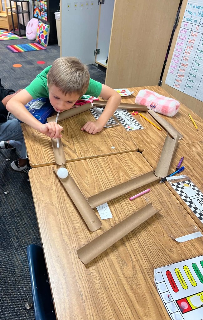 A child uses a straw to guide a ball through a maze of cardboard tubes on a table.