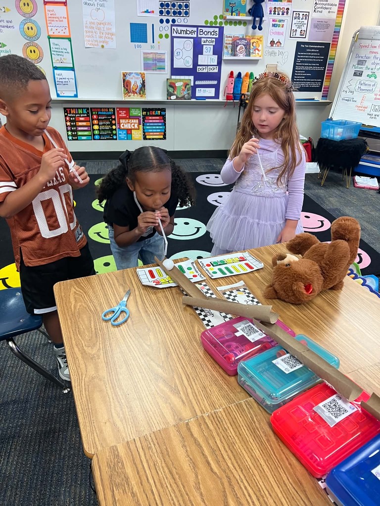 Three children engage in a classroom activity. One boy holds a pipette, another girl uses a pipette, and a teddy bear sits on the table.