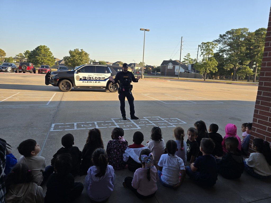 Police officer stands on hopscotch pattern in parking lot, students sit in semicircle in front of him