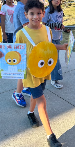 A boy in a yellow shirt with a cheese-shaped backpack walks with books and smiles at camera. Behind him are other students in casual clothing.