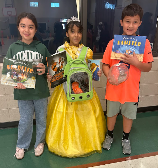 Three children stand inside with books. One wears a yellow dress, one a green sweatshirt, and one an orange shirt.