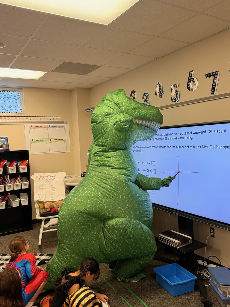 Person in dinosaur costume teaching class, kids seated on floor, large screen behind them with text and numbers.