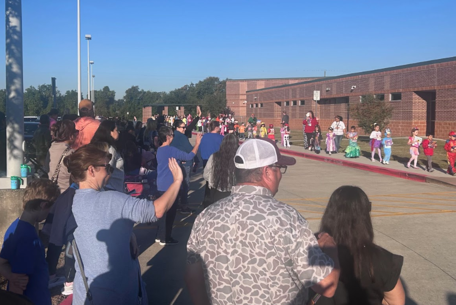 Crowd of people standing outdoors near a school building, some in costumes, on a clear day.