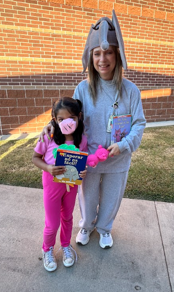 Two children wearing costumes, one holding a book, stand on a sidewalk in front of a brick wall.