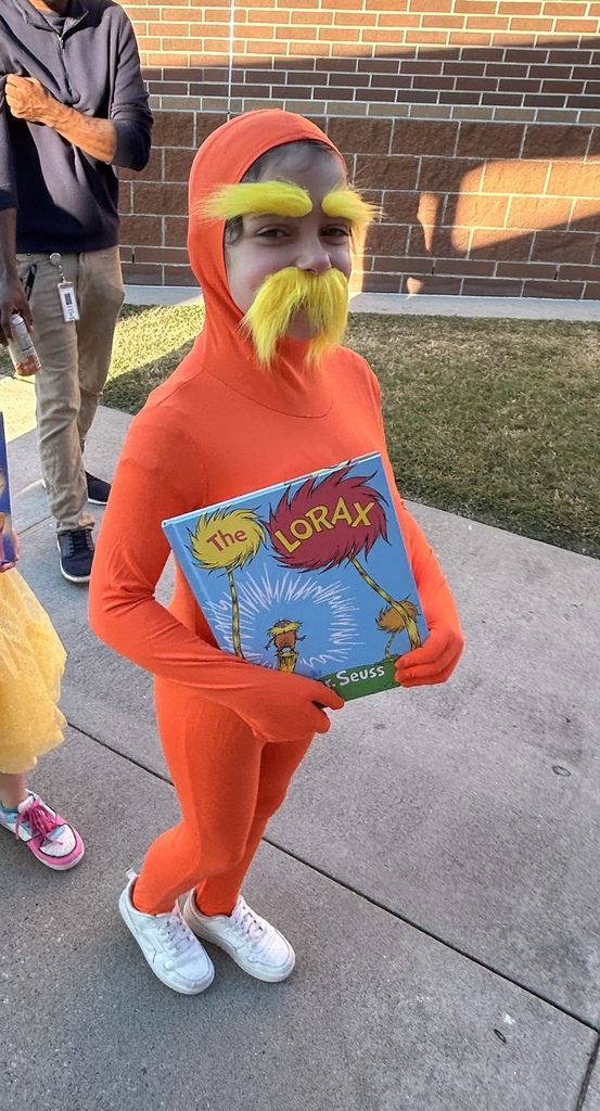 A young person dressed in orange holds a book. A brick wall and a person wearing sneakers are behind them.