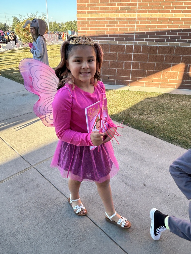 A girl in a pink fairy costume stands on a sidewalk, smiling and holding a book.