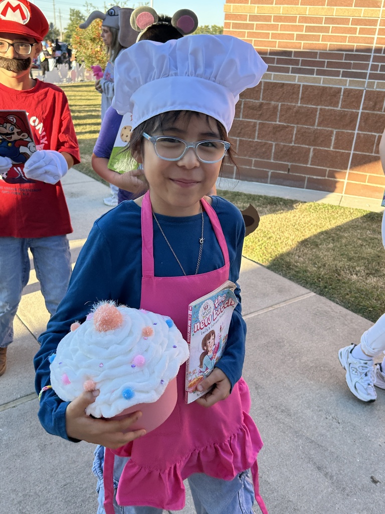 Child in costume holding a cupcake and book outside school with brick wall behind.