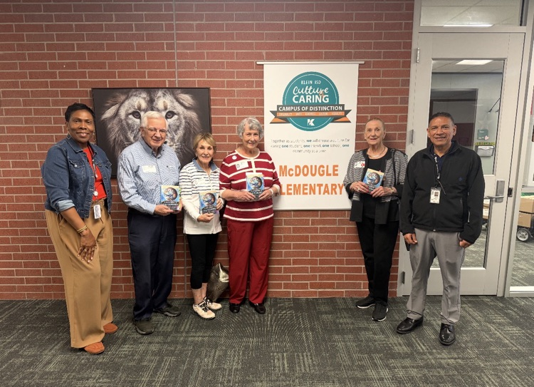 Group of adults standing in front of a brick wall. One person holds a book.