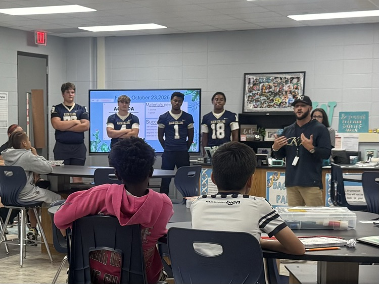Group of people in football jerseys standing by a monitor, others seated at tables.