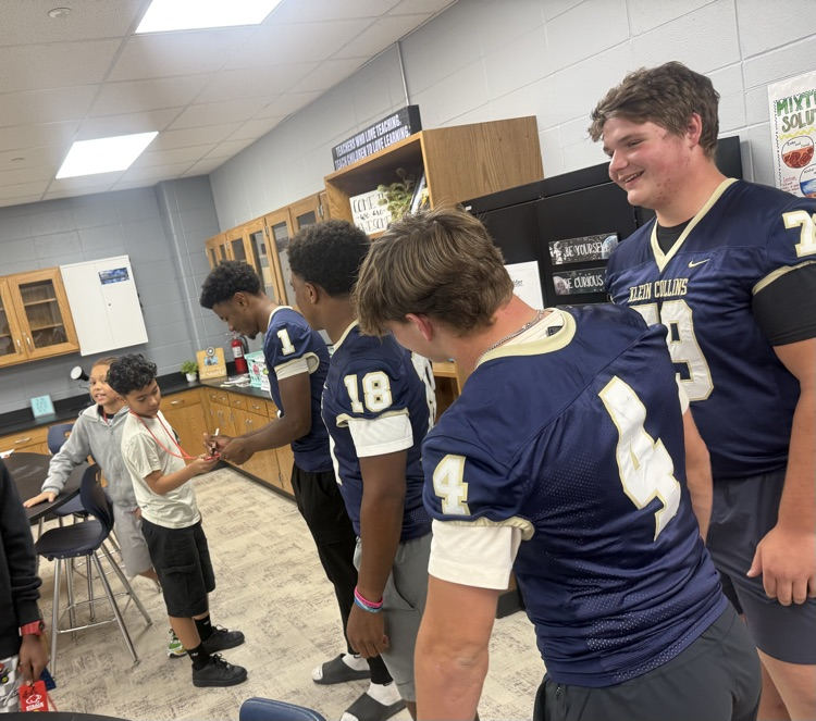 High school football players wearing blue jerseys and shorts stand in a room, talking to kids.