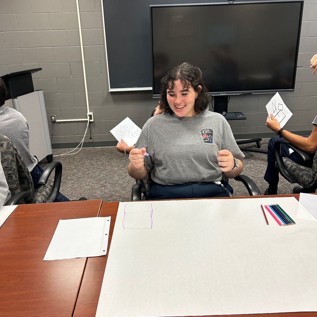 Student drawing on large paper while sitting back to back with another student who is describing the photo