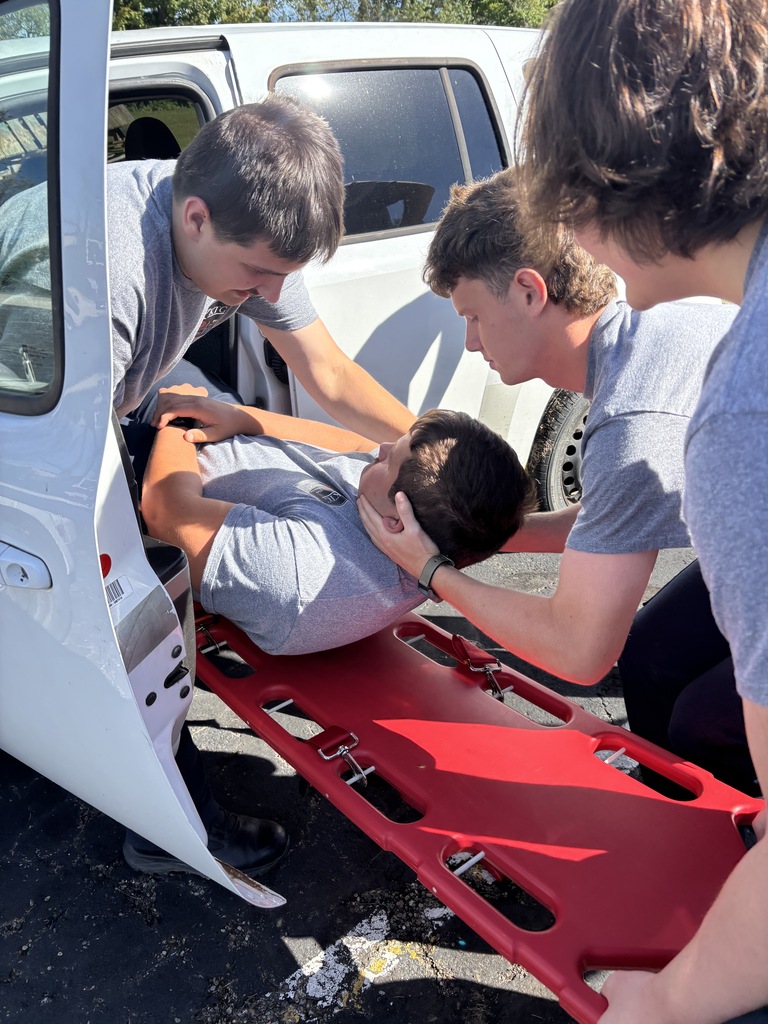 Close-up of EMS trainees practicing C-spine stabilization while sliding a patient onto a red backboard out of a truck door.