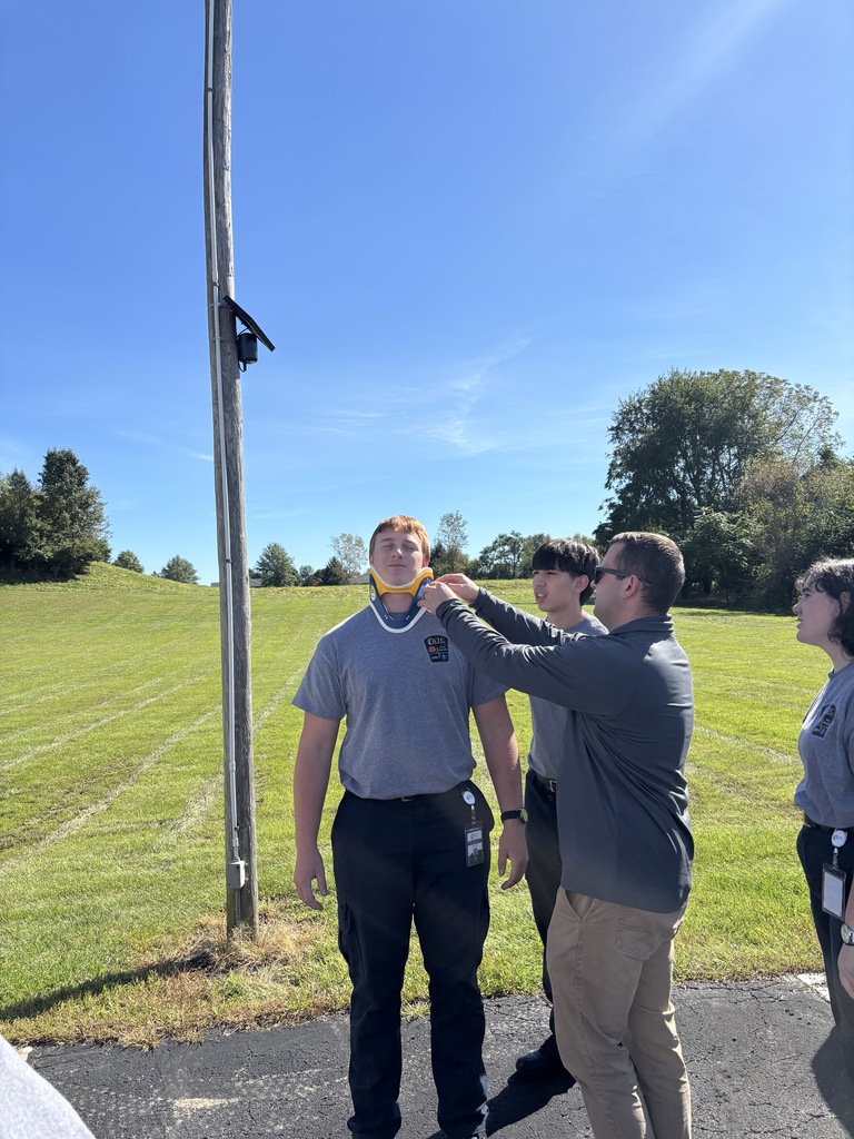 An instructor and student adjust a cervical collar on a classmate during an outdoor EMS training exercise.