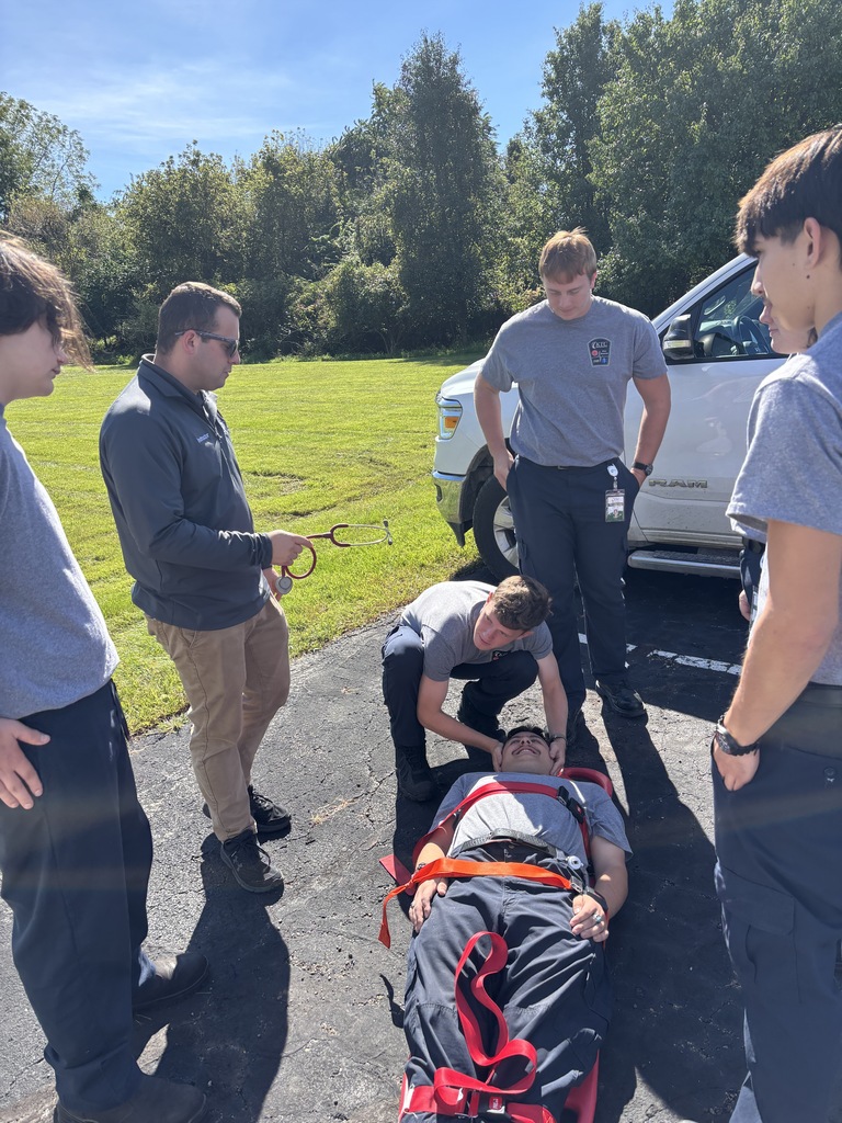EMS instructor with a stethoscope guiding trainees as they practice securing a patient on a spine board outdoors.