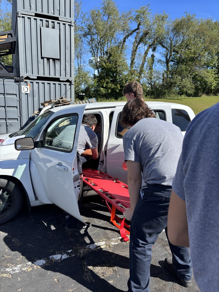 EMS trainees positioning a red spine board into the open door of a white SUV during a vehicle extrication exercise.