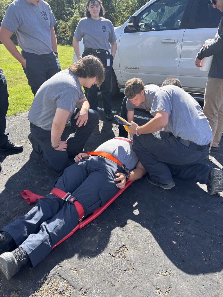 EMS students or trainees practicing immobilizing a patient on a red backboard outdoors near a white car.